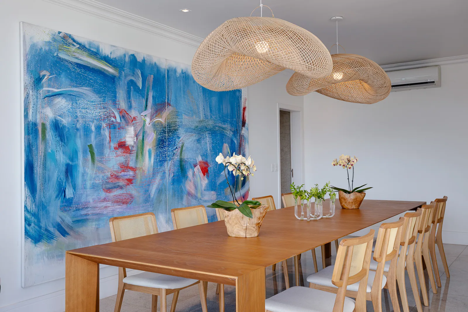 Long wood dining table under woven pendant lights, blue canvas behind